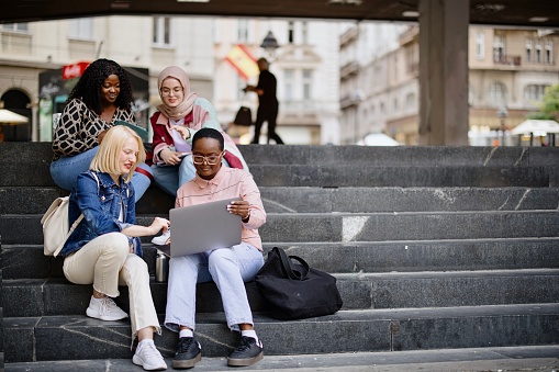 Four multiethnic girl friends walk around the city after or before the lecture. They wear casual clothes, holding electronic devices such as laptops, digital tablets or smartphones. They discuss lectures, talk and laugh, look at funny memes.