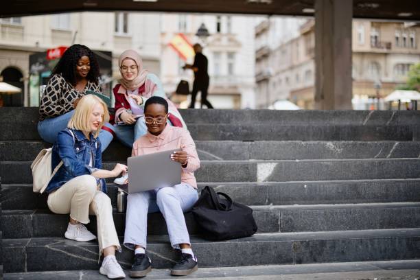 Students sit on the steps near the college and look at the laptop and  digital tablet and talk stock photo