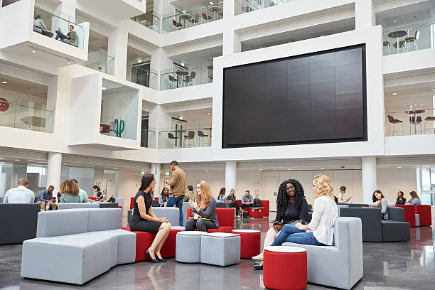 students sit talking under av screen in atrium at university - lobby stockfoto's en -beelden