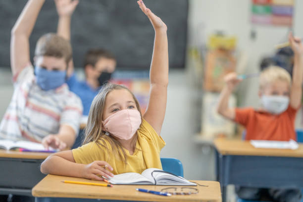 Students wearing masks in class Group of students wearing protective face masks while raising their hands in class. primary school stock pictures, royalty-free photos & images