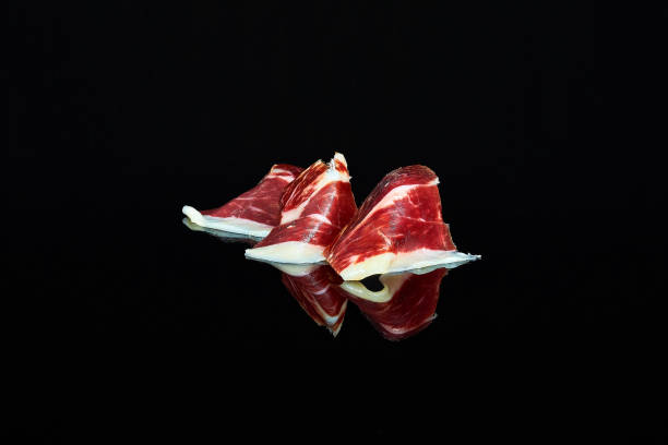studio photo of a series of slices of 100% Iberian acorn-fed ham folded on black background with reflection. stock photo