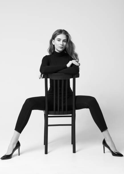 Studio portrait of beautiful skinny girl. Pretty young woman dressed in turtleneck sweater, leggings and shoes sitting on stool. Black and white stock photo