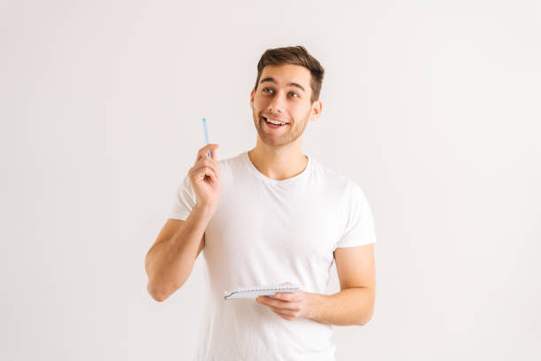 Studio portrait of enthusiastic young man writing in copybook, raising pen up eureka gesture on white isolated background. stock photo