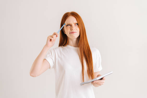 Studio portrait of pondering redhead young woman scratches head with pencil, holding diary looking down, standing on white isolated background. stock photo