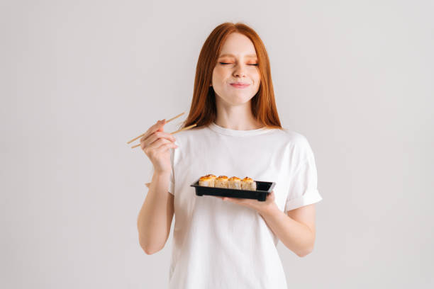 Studio portrait of satisfied young woman with closed eyes eating delicious sushi rolls with chopsticks standing on white isolated background. stock photo