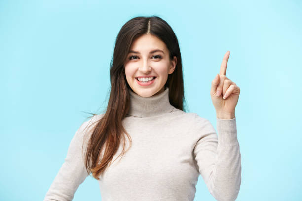 studio portrait of young caucasian woman beautiful young caucasian woman showing a number one sign, looking at camera smiling, isolated on blue background number 1 stock pictures, royalty-free photos & images