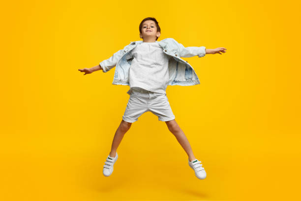 Stylish boy leaping up and stretching out arms Funny youngster in trendy outfit stretching out arms and jumping against yellow background boy jumping stock pictures, royalty-free photos & images