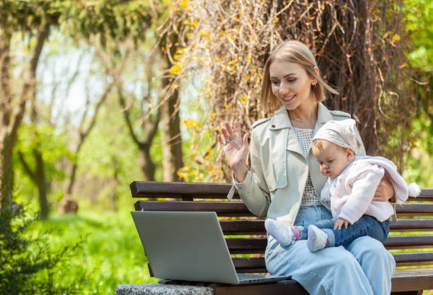 Stylish business mom makes a video call with a laptop and her daughter in her arms while sitting on a bench in the park. Distance work, motherhood, work on maternity leave stock photo