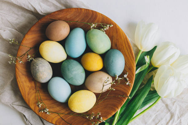 Stylish Easter eggs in wooden plate, tulips and linen napkin on rustic white table, flat lay. Happy Easter! Natural dyed colorful eggs and spring flowers rustic composition. stock photo
