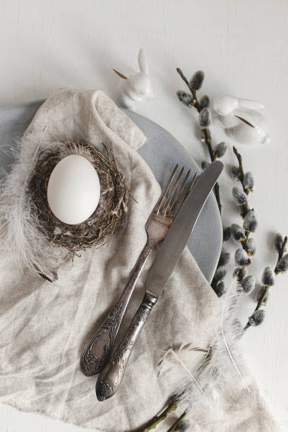 Stylish Easter table setting flat lay. Natural easter egg in nest, pussy willow branches, feathers on modern plate with napkin and cutlery on white wooden table. Modern Easter table decoration stock photo