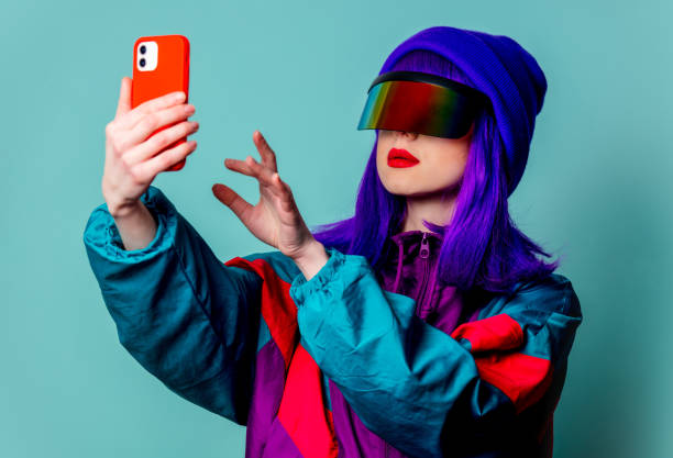 Stylish girl in cyber punk glasses and 80s tracksuit using mobile phone on blue background stock photo
