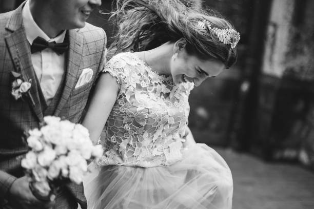 Stylish happy bride and groom walking on background of old church in rain, close up. Provence wedding. Beautiful emotional wedding couple smiling. Romantic moment, black and white image stock photo