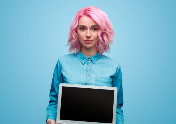 Stylish hipster woman showing laptop Modern young woman in blue shirt and with pink hair holding modern laptop on blue background looking at camera pink hair stock pictures, royalty-free photos & images