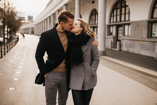 A girlfriend and boyfriend very much in love are kissing while strolling along a beautiful colonnade. They have their eyes closed. She is wearing a trendy gray winter coat and a warm black scarf. He is dressed in a fashionable wollen brown sweater with a black coat over it. Horizontal outdoor daylight photo.