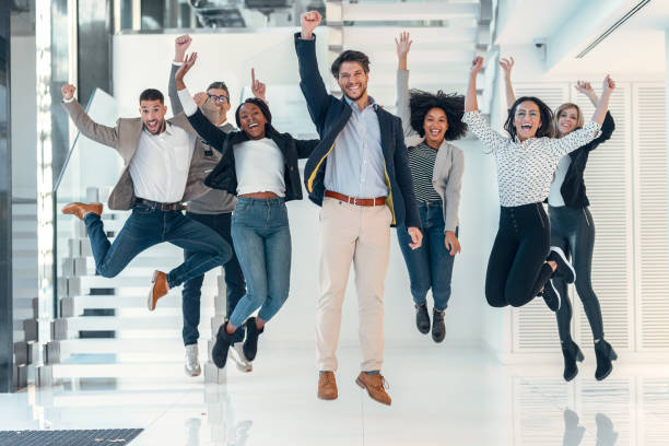 Successful business multiage team jumping while celebrating successes while looking at camera in modern startup. stock photo