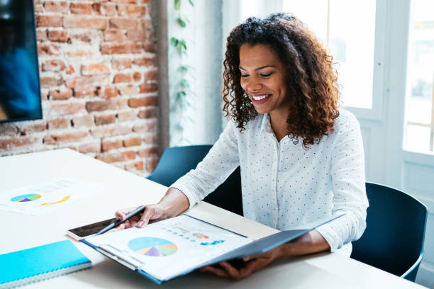 Successful businesswoman analyzing financial reports at work. Elegant african-american businesswoman sitting in her office holding paper documents containing graphs and financial data. report document stock pictures, royalty-free photos & images