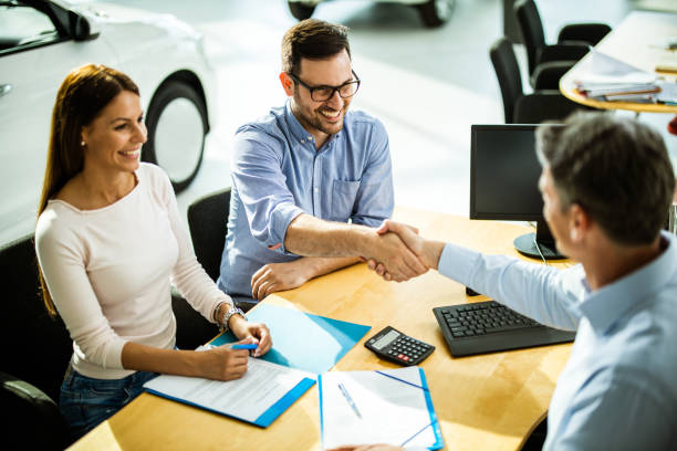 Successful deal in a car showroom! Happy couple buying a car in a showroom while men are shaking hands after reaching a successful deal. Focus is on young man. car salesperson stock pictures, royalty-free photos & images