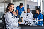 Portrait of mature female doctor sitting in meeting room with specialist and nurses discussing case in background. Successful woman doctor in labcoat and stethoscope in hospital. Smiling pediatrician looking at camera with medical staff brainstorming in background.