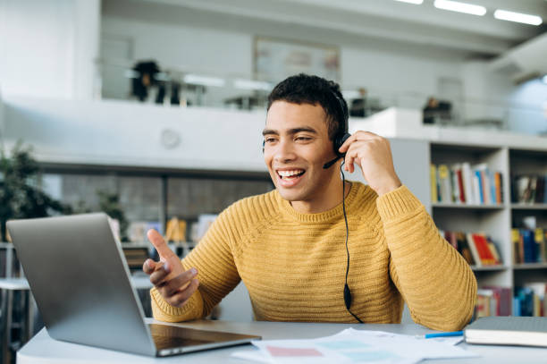 Successful hispanic freelancer, manager or tutor sitting at work desk wearing headphones communicate and smiling while video conference with colleagues or students stock photo