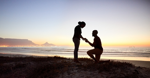 Silhouette of young man making proposal wearing engagement ring to his girlfriend at the beach in evening