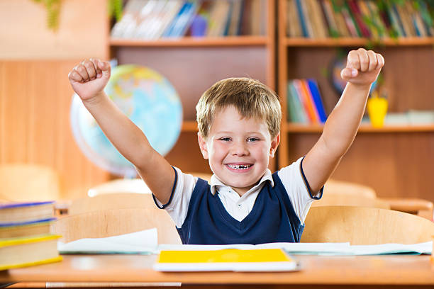 Successful schoolboy with hands up sitting at desk stock photo