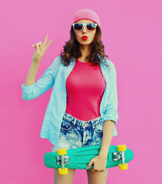 Summer colorful portrait of stylish modern young woman model posing with green skateboard and blowing her lips wearing a pink hat on background stock photo