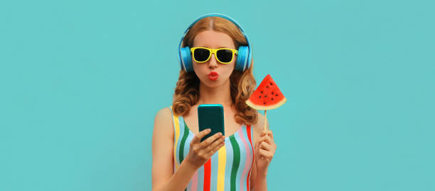 Summer colorful portrait of stylish young woman in headphones listening to music on smartphone with juicy lollipop or ice cream shaped slice of watermelon on blue background stock photo