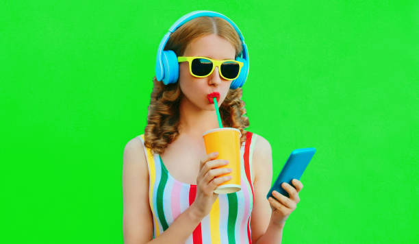 Summer colorful portrait of stylish young woman listening to music in headphones looking at smartphone and drinking fresh juice on green background stock photo