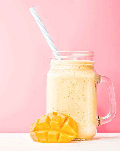 summer drink mango milkshake in mason jar with drinking straw decorated with a half of mango on blue background summer drink mango milkshake in mason jar with drinking straw on pink background mango smoothie stock pictures, royalty-free photos & images