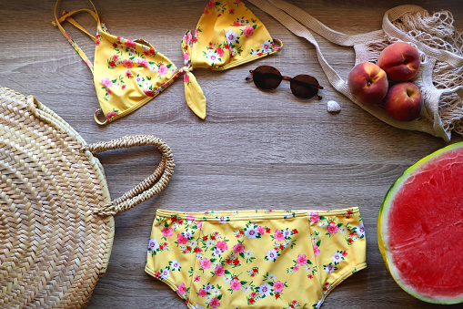 Yellow floral swimming suit, sunglasses, mesh tote with fresh peaches, seashell, watermelon half and wicker bag on wooden background. Summer essentials, flat lay.