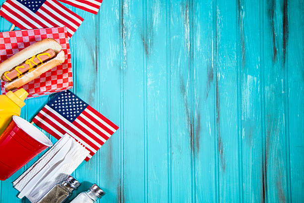 Summer holiday picnic. Hot dogs, USA flags. Blue wooden table. Happy Independence Day America!  Left border of: USA flags, hot dog, cup, mustard, utensils, salt and pepper shakers.  Copyspace to right. Teal blue wooden beadboard background with grunge painted effects.  Memorial Day, July 4th, Veteran's Day, Labor Day, Flag Day summer picnic concepts.  American pride and patriotism. memorial day background stock pictures, royalty-free photos & images