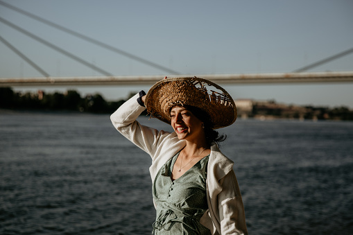 Cheerful casual dressed female with a hat on a head enjoying a sunny windy day by the water/river. Vacation and lifestyle concept