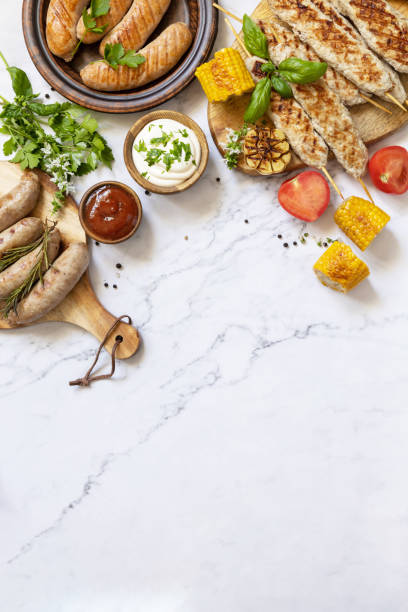 Summer party food. Grill food menu. Various bbq grilled sausages with fresh herbs and spices on marble tabletop. View from above. Copy space. stock photo