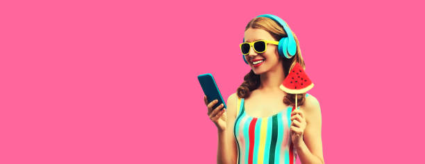 Summer portrait of happy smiling young woman model in headphones listening to music on smartphone with juicy lollipop or ice cream shaped slice of watermelon on pink background stock photo