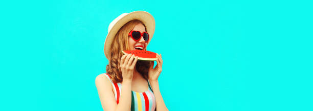 Summer portrait of happy young woman eating fresh tasty slice of watermelon wearing straw hat, red heart shaped sunglasses on blue background, blank copy space for advertising text stock photo