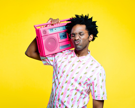 Summer portrait of afro american young man with dreadlocks wearing vintage shirt with lightning pattern and gold glasses, holding pink boom box on his shoulder. Studio shot on yellow background.
