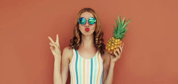 Summer portrait young woman blowing her lips with pineapple wearing a sunglasses posing on a brown background stock photo