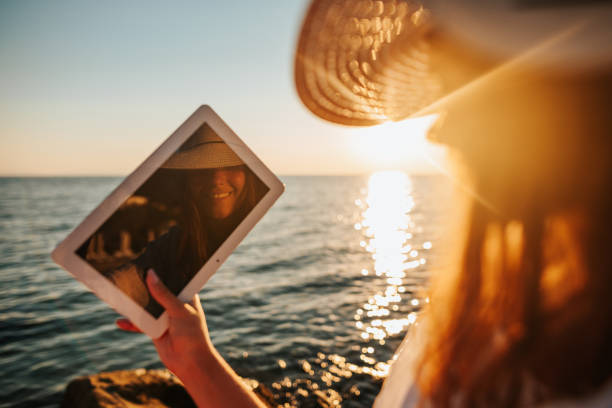 Summer selfie Beautiful young woman in white sun dress and with hat taking selfie with her digital tablet on the beach at beautiful summer sunset above the sea summer girl stock pictures, royalty-free photos & images