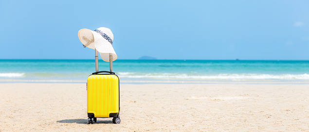 Summer traveling and tourism planning with yellow suitcase luggage with big hat fashion in the sand beach. Travel in the holiday, blue sky and beach background. Summer and Travel Concept, copy space for banner