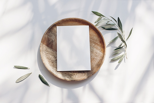 Summer wedding stationery mock-up scene. Blank greeting card, wooden plate, olive tree leaves and branches in sunlight, white table background with palm shadows. Feminine flat lay, top view.