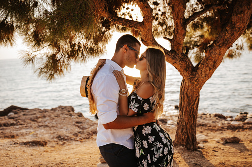 Loving young couple standing and kissing by the tree on coastline at summer sunset, side view