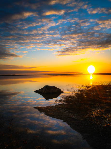 Sunrise, cumulous clouds, beach grass, black rock, reflections in the water, white sun, orange horizon, and blue sky. Tranquil zen-like seascape on Cape Cod. stock photo
