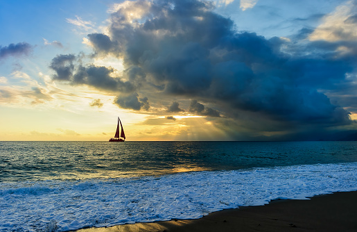 A Sailboat Silhouette Is Sailing Towards The Sun Rays Emanating From The Sunset Sky