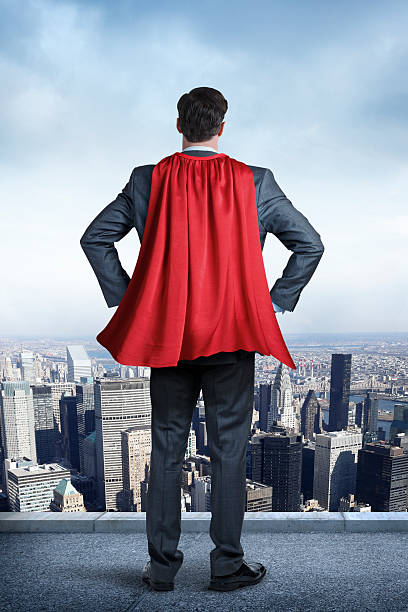 Superhero Businessman Wearing Red Cape Looking At Big City A businessman wearing a red cape looking out over the city below him. His hands are on his hips.  Blue sky and patchy clouds are in the distance. cape garment stock pictures, royalty-free photos & images