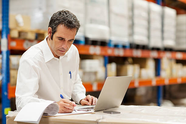 Supervisor working in warehouse Supervisor doing paperwork while using laptop in warehouse supply chain management stock pictures, royalty-free photos & images