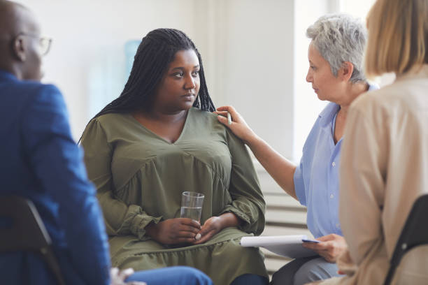 Support Group Circle Portrait of sad African-American woman listening to psychologist during support group meeting with people siting in circle and comforting her mental health awareness stock pictures, royalty-free photos & images