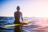 surfer girl sitting on surfboard in ocean, sunset.