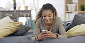 A mixed race teenage girl is enjoying time on her smart phone in her bedroom. Here, she listens to music with headphones.