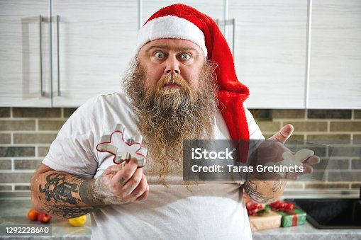 istock A surprised bearded with tattooed arms man wearing like Santa Claus holding person-shaped piece of dough on one hand and a metal man-shaped cookie-cutter on the other 1292228640