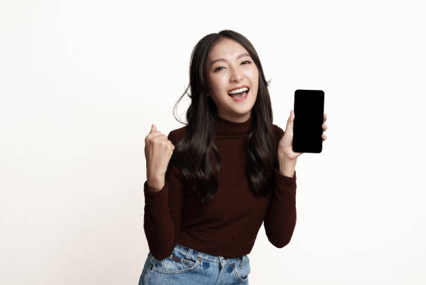 überraschte junge asiatische frau, die auf weißem hintergrund mit smartphone steht. - weiblicher teenager fotos stock-fotos und bilder